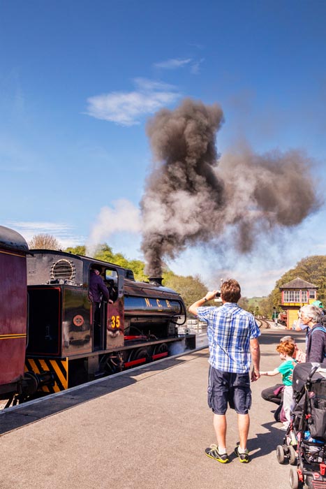Family watch and take photos at Bolton Abbey Station, as 0-6-0 Austerity Class Saddle Tank Engine blows steam as it leaves the station. Yorkshire Dales National Park, North Yorkshire, England, UK