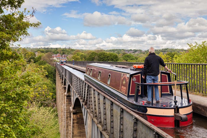 Man with his hand on the tiller, steering narrowboat over the Pontcysyllte Aqueduct, built by Thomas Telford, on the Llangollen branch of the Shropshire Union Canal, near Llangollen, Denbighshire, Wales, UK