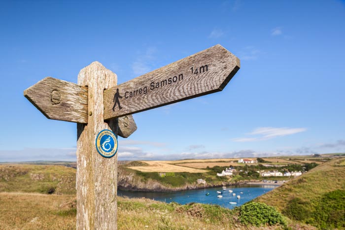 Signpost showing a footpath to Carreg Samson, a Neolithic dolmen grave on the Pembrokeshire coast of Wales, with Abercastle village and harbour in the background.