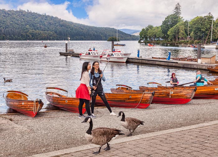 Two Asian girls taking photos with a selfie stick on the shores of Lake Windermere, Cumbria, England, UK